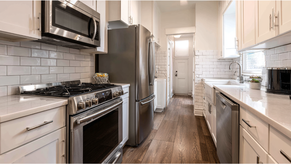 A narrow galley kitchen with white cabinetry, stainless steel appliances, subway tile backsplash, and dark wood flooring.