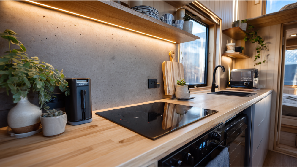 A modern compact kitchen with light wood countertops, concrete backsplash, and open shelving, featuring potted plants and under-cabinet lighting.